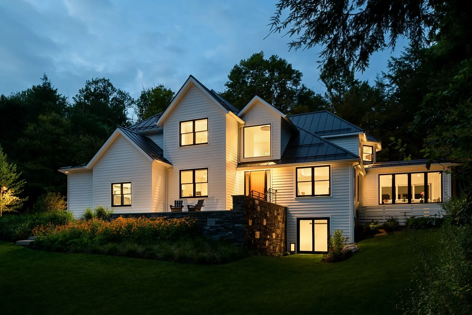 A white Vermont farmhouse at dusk with warm light in the windows and a lit stone wall